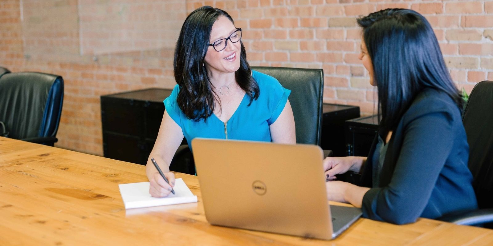 One lady working on a pad with pen next to another working on a laptop at a desk